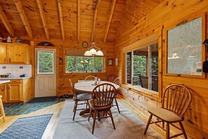A wooden cabin interior with a dining area, kitchen, and large windows.