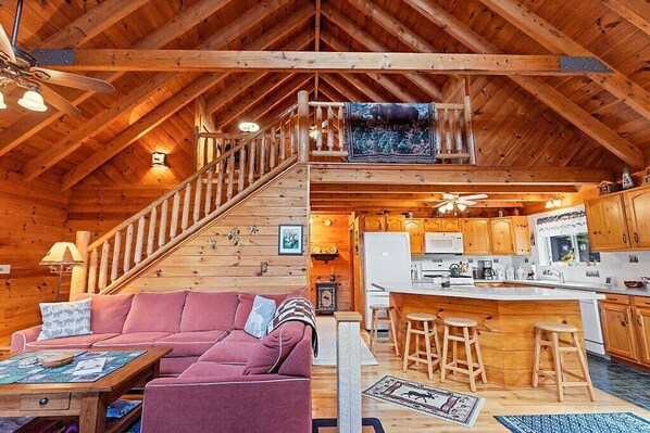 A cozy log cabin interior with a living room, kitchen, and a staircase leading to an upper level.