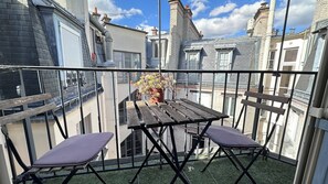 Ein Balkon mit Holztisch und zwei gepolsterten Stühlen, einem Blumentopf und Blick auf angrenzende Gebäude.