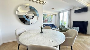 A modern living room with a round marble table, comfortable seating, a large abstract wall art piece, and a view of the outdoors through large windows. - Luxury Apartment – 3 Bedrooms / Arc de Triomphe (Paris)
