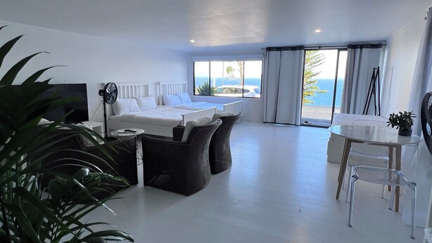 A modern bedroom with a bed, a small table, and a chair. There is a large window with curtains, and a view of the outdoors. - Ocean View In Clifton studio (Clifton)