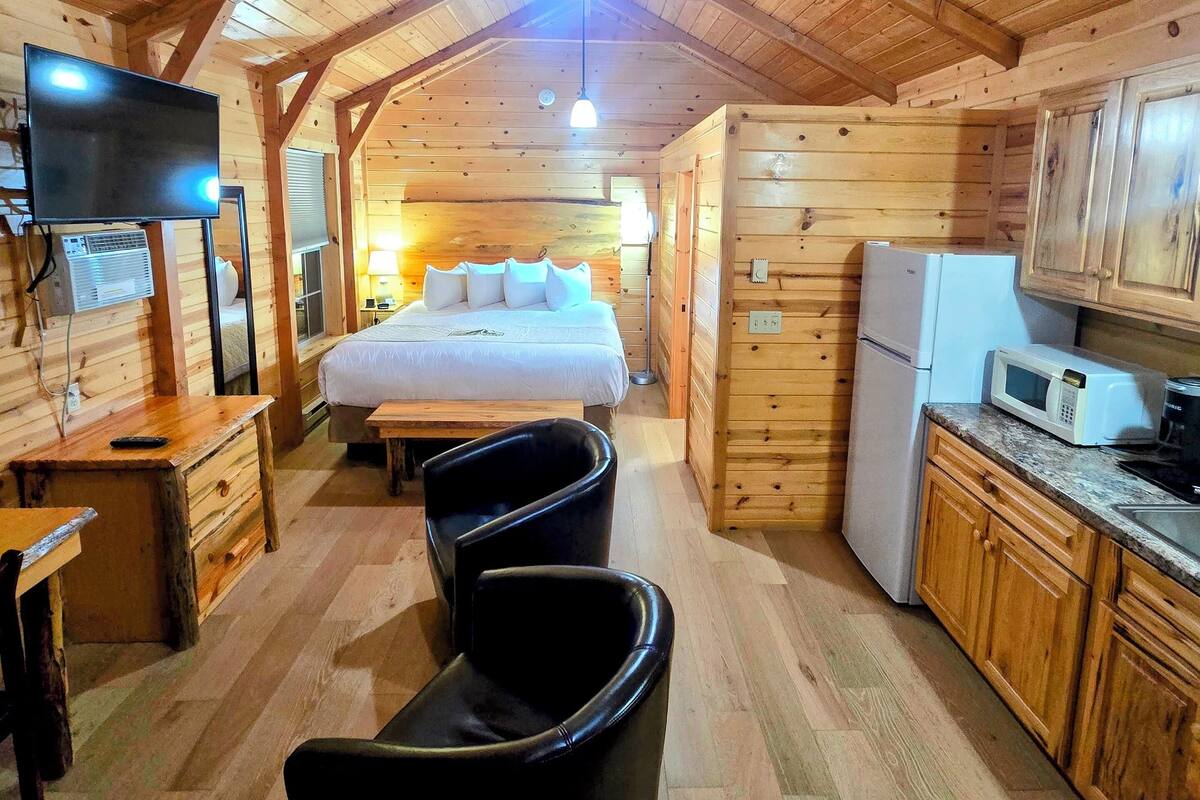 A cozy cabin-style room with a bed, a desk, a chair, a kitchen area, and a television.