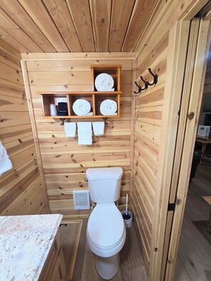 A wooden cabin bathroom with a toilet, a shelf with towels, and a small table.