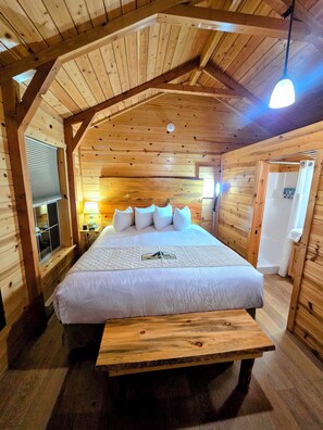 A wooden cabin bedroom with a large bed, a bench, a window with blinds, and a wooden ceiling.
