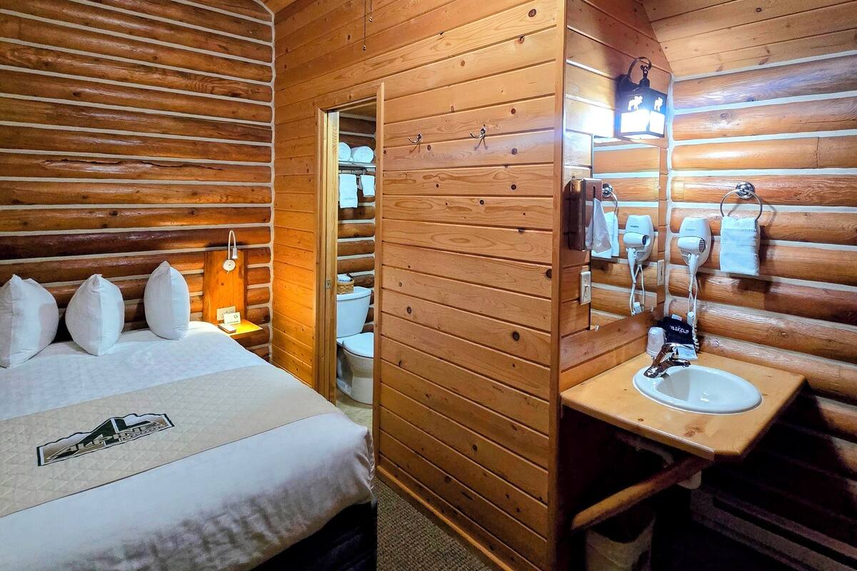 A wooden cabin-style room with a bed, a ceiling fan, a bathroom with a sink and mirror, and a toilet.