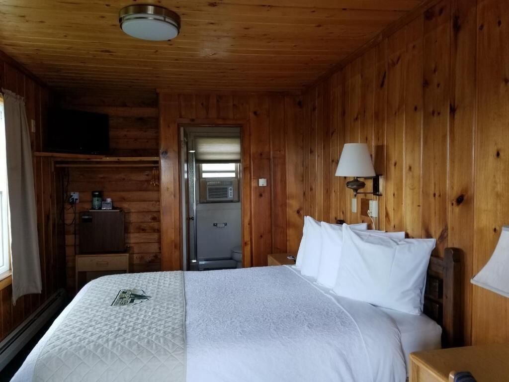 A wooden cabin bedroom with a bed, bedside lamps, and a door leading to a bathroom.