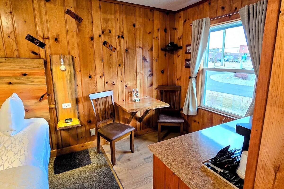 A cozy cabin-style room with wooden walls, a bed, a dining table, and a small kitchen area.
