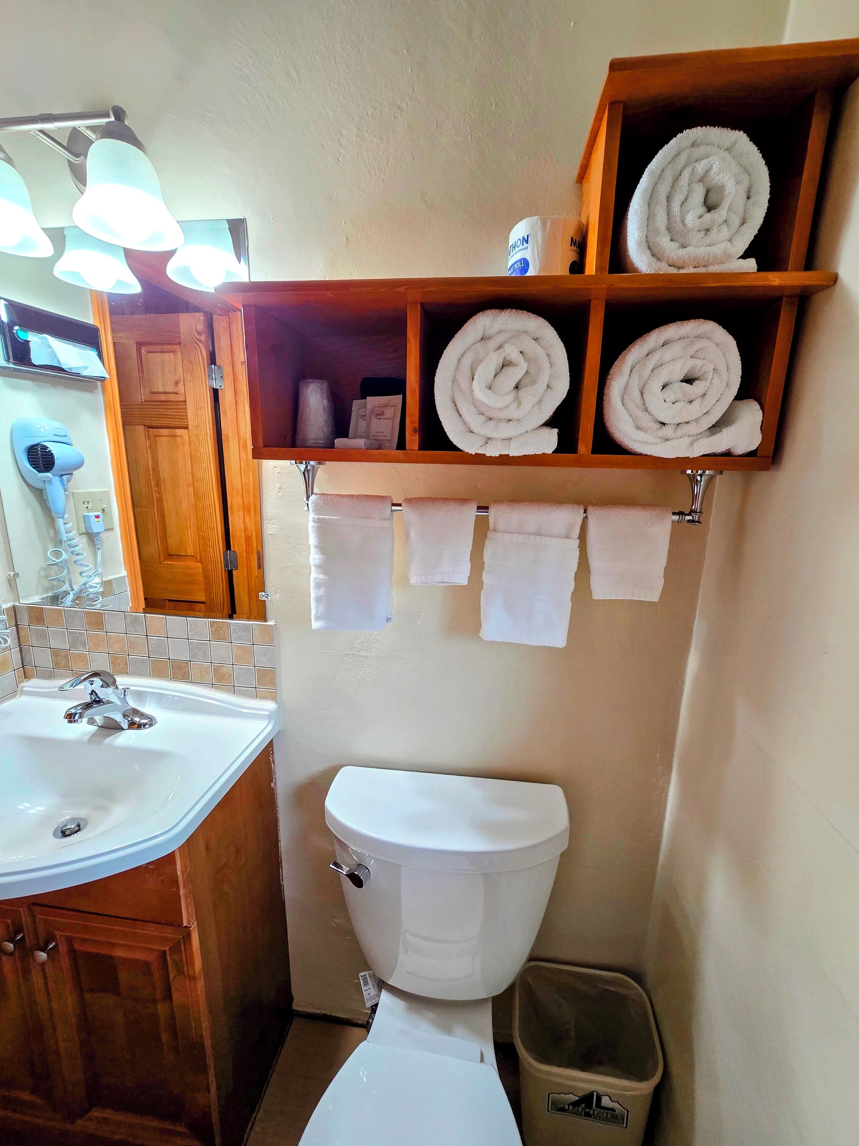 Une petite salle de bain avec un lavabo blanc, des toilettes et une étagère au-dessus des toilettes où sont rangées des serviettes pliées.
