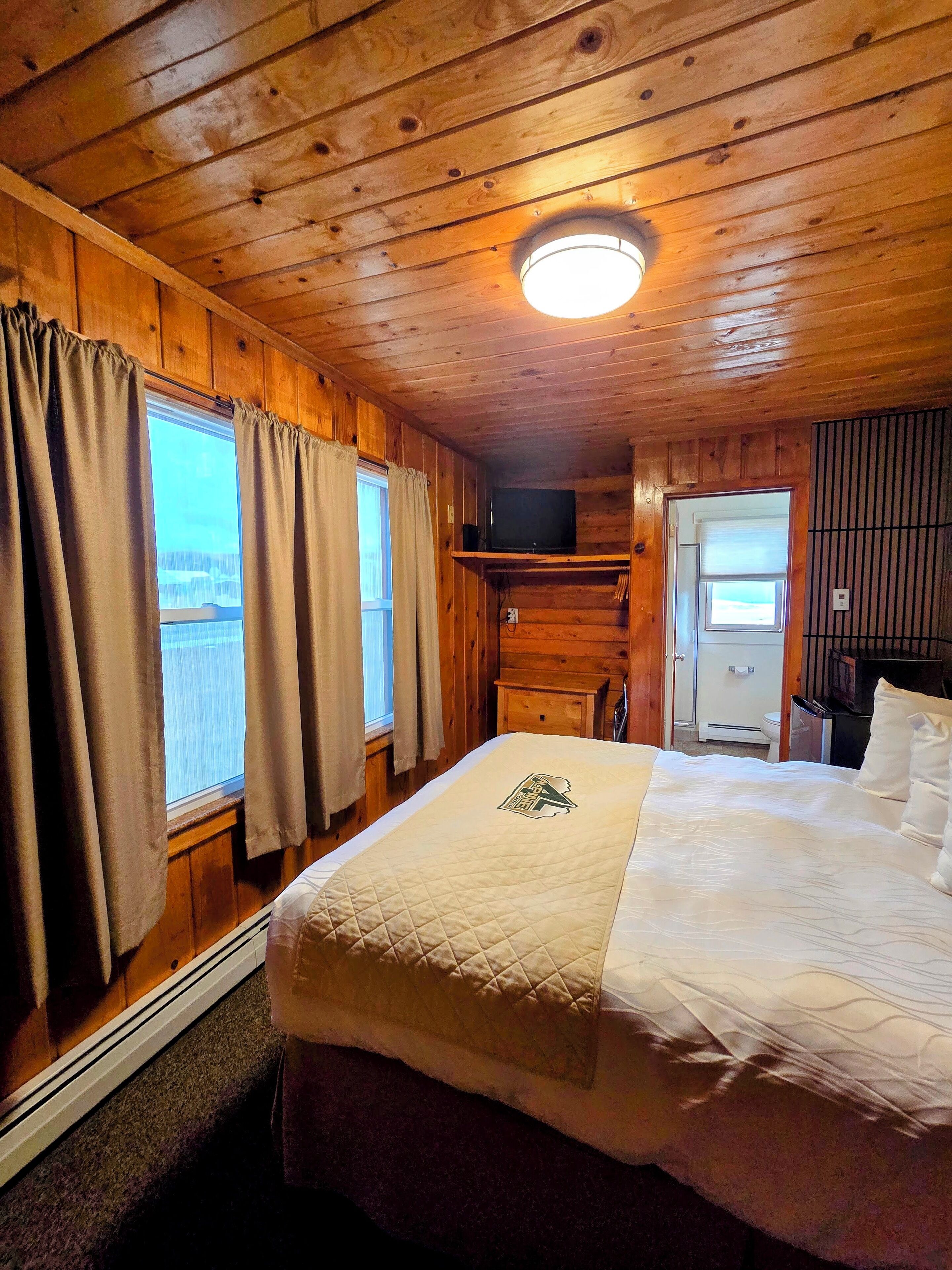 Una habitación de cabaña de madera con una cama, un televisor en una estantería y una ventana con cortinas.