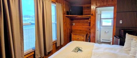 Una habitación de cabaña de madera con una cama, un televisor en una estantería y una ventana con cortinas.