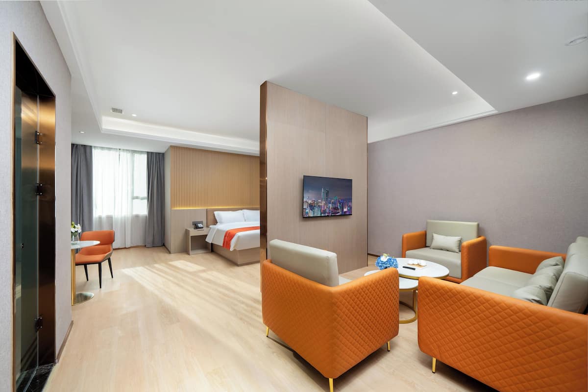 A hotel room with a bed, two orange armchairs, a white sofa, a small round table, and a painting on the wall.