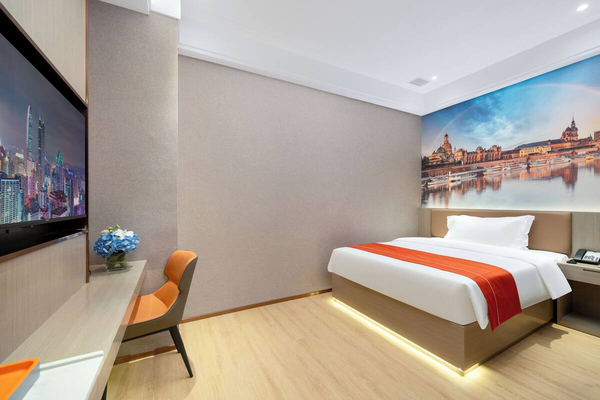 A hotel room with a bed, a desk, and a chair. There is a painting on the wall and a TV mounted on the wall.