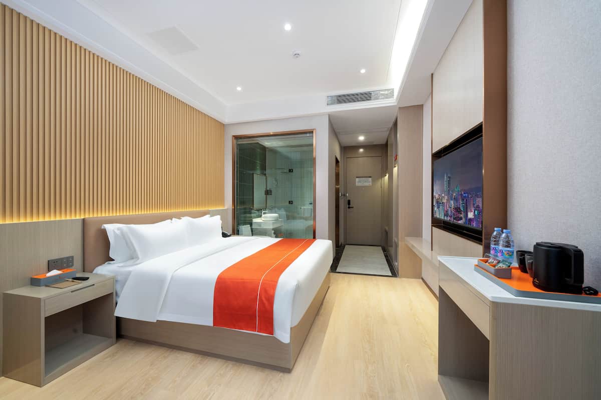 A modern hotel room with a bed, bedside table, TV, and a view of the cityscape.