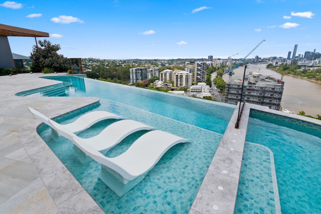 Une piscine à débordement située sur le toit, avec des chaises longues blanches, une vue sur la ville et une grue en arrière-plan.