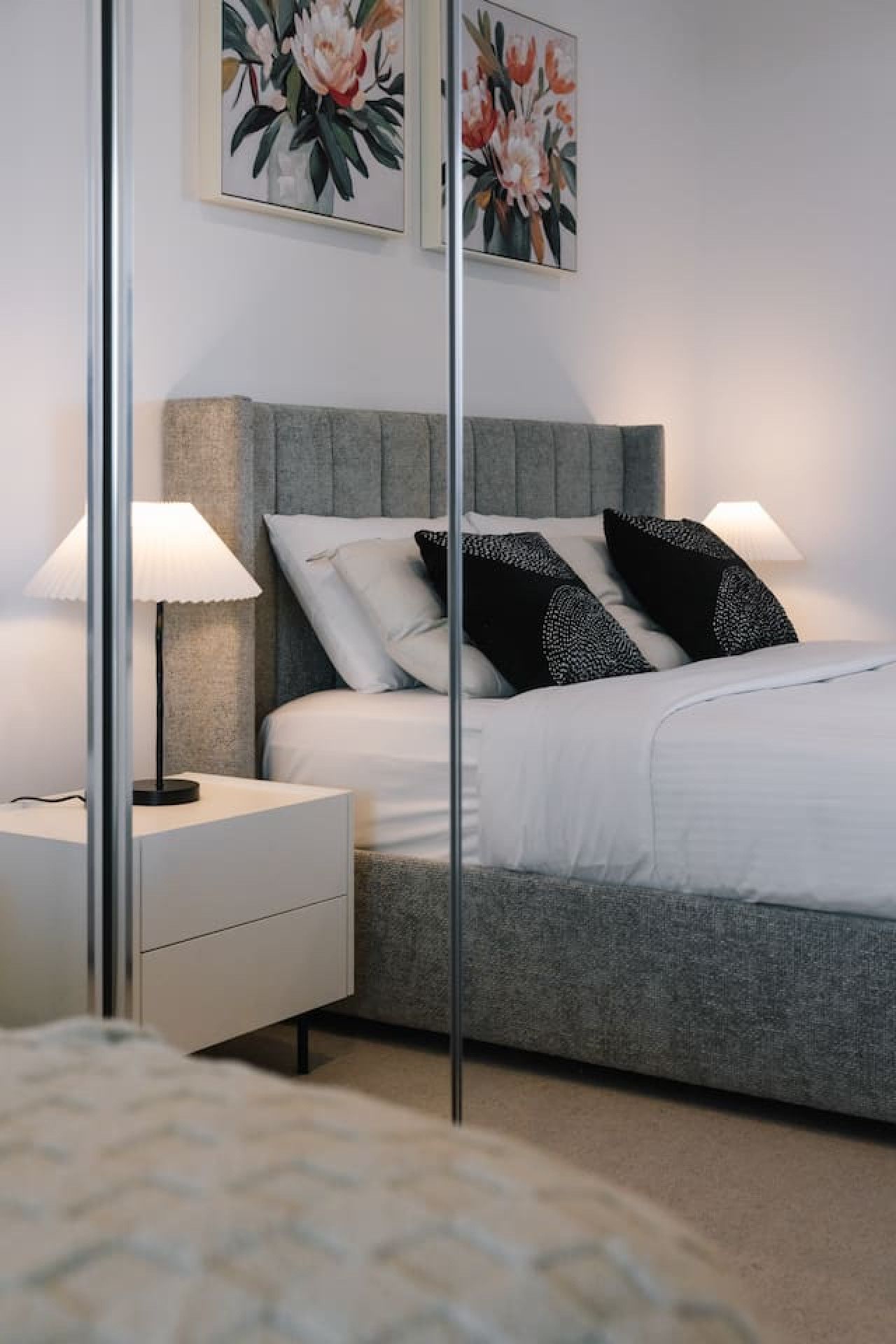 A bedroom with a grey upholstered headboard, white bedding, two black patterned pillows, and a bedside table with a lamp.
