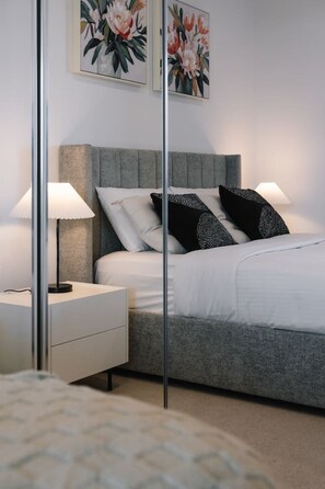 Ein Schlafzimmer mit grauem, gepolstertem Kopfteil, weißer Bettwäsche, zwei schwarzen Kissen mit Muster und einem Nachttisch mit Lampe.