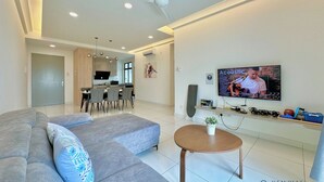 A modern living room with a grey sofa, a coffee table, and a flat-screen TV mounted on the wall. There is a kitchen area in the background with a dining table and chairs. - The Platino Paradigm Mall by Stayrene (Johor Bahru)