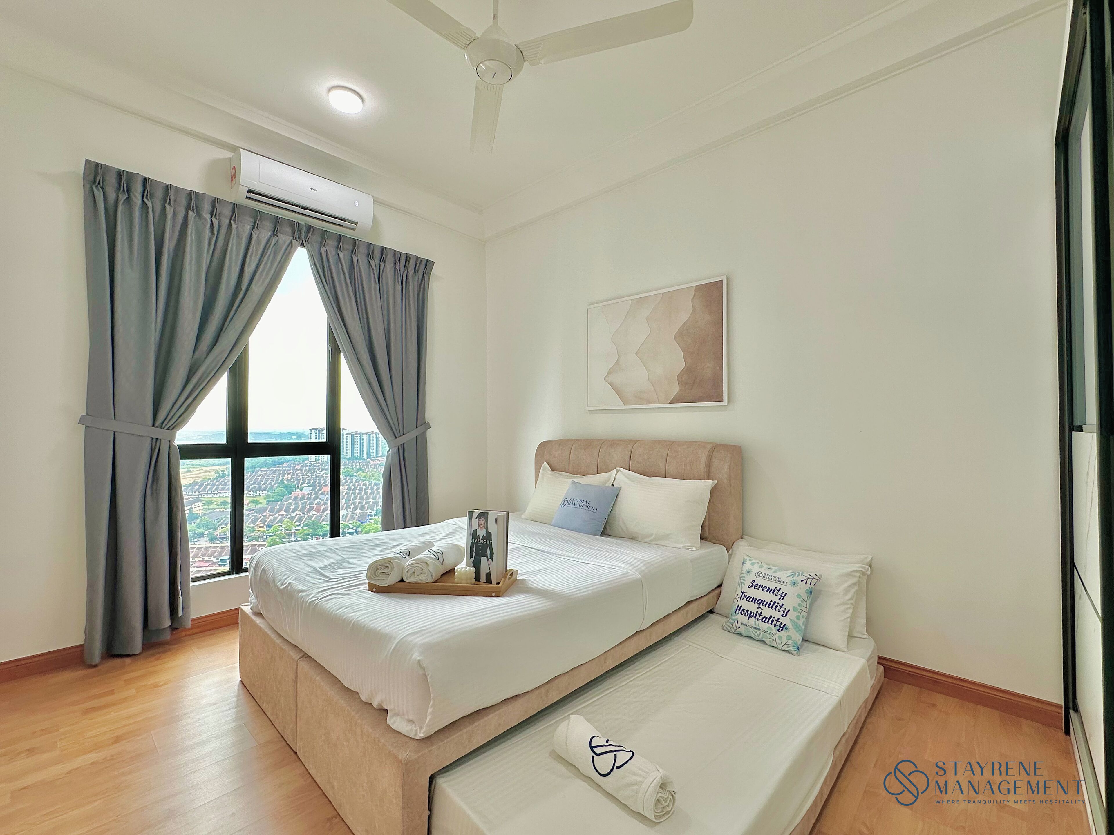 A hotel room with a bed, a ceiling fan, an air conditioning unit, and a view of the city through large windows.