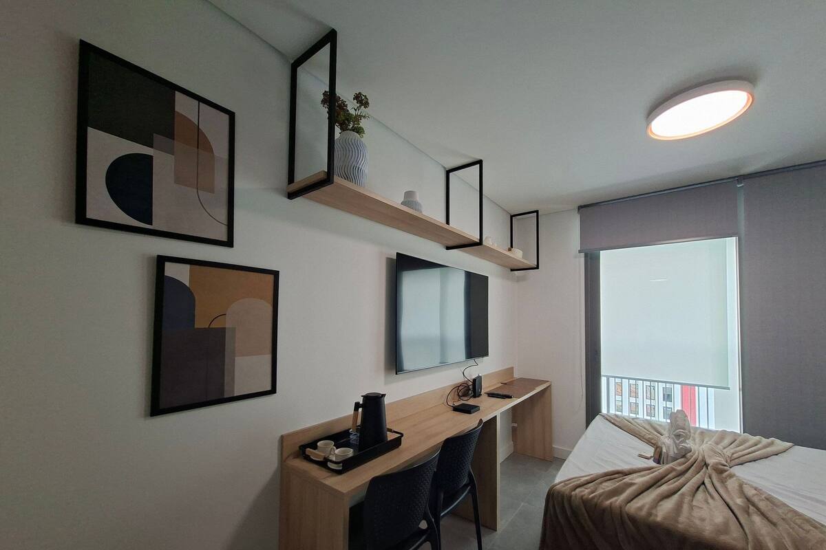 A modern hotel room with a bed, a desk with a chair, a flat-screen TV, and artwork on the walls.