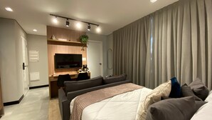 Una habitación de hotel moderna con cama, escritorio, televisor y un ventanal con cortinas.