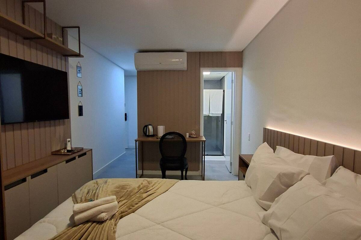 A hotel room with a bed, a desk, a chair, a television, and an air conditioning unit.