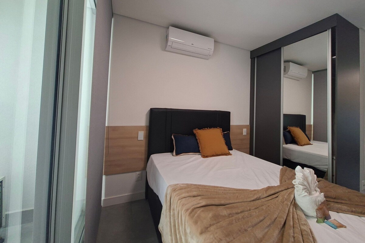 A hotel room with a bed, a large mirror, an air conditioning unit, and a window with curtains.