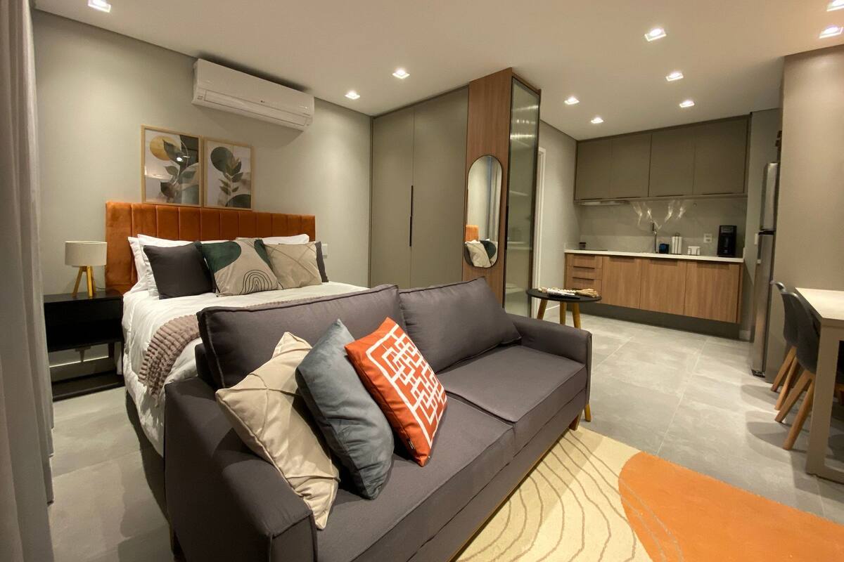 A modern bedroom with a bed, a sofa, a small table, and a kitchen area.