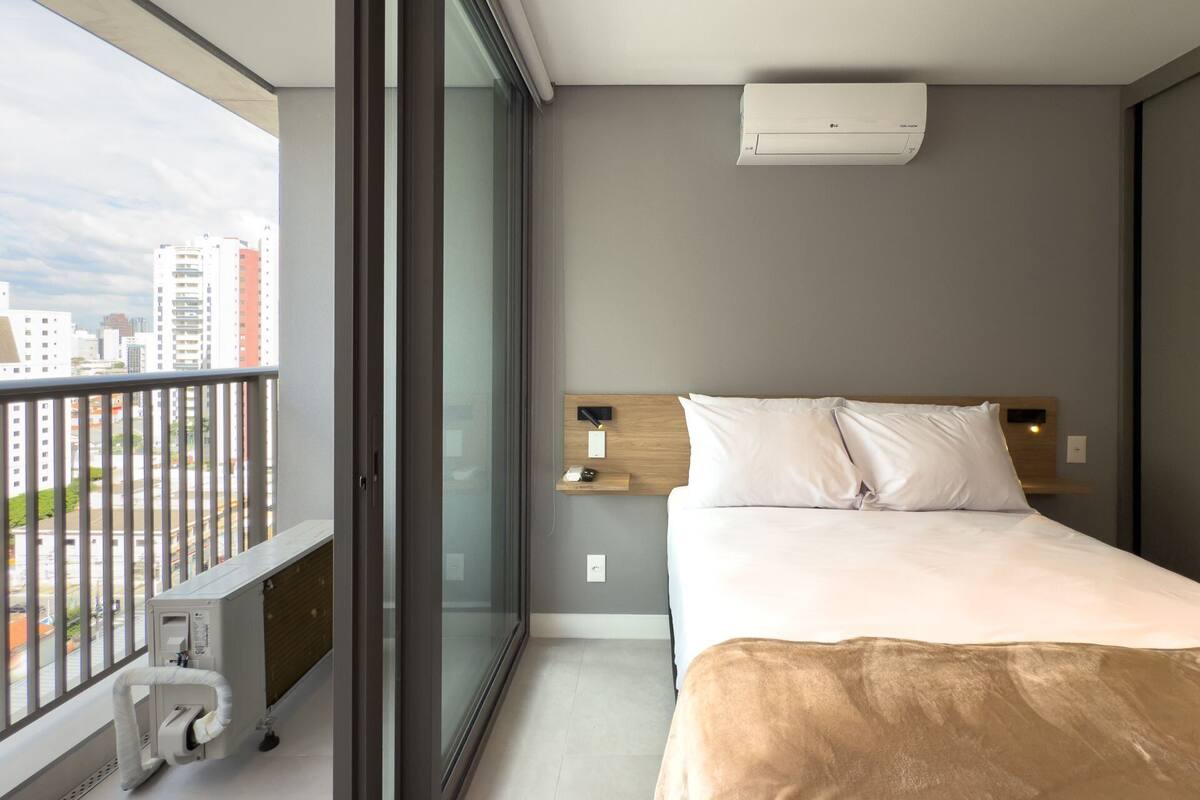 A modern hotel room with a large window, a bed with white bedding, a wooden headboard, a bedside table with a lamp, and an air conditioning unit on the wall.