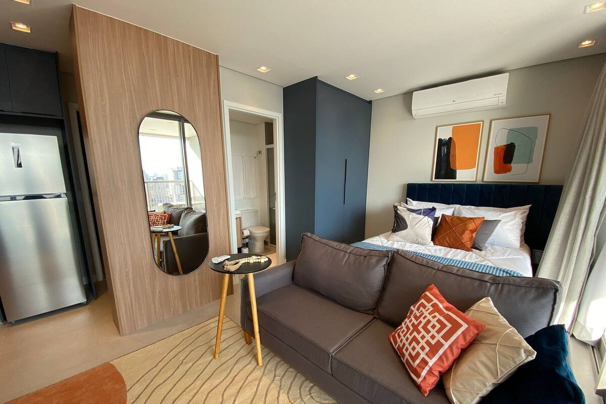 A modern hotel room with a sofa, a small table, a bed with pillows, a mirror, and an air conditioning unit.