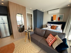 A modern hotel room with a sofa, a small table, a bed with pillows, a mirror, and an air conditioning unit.