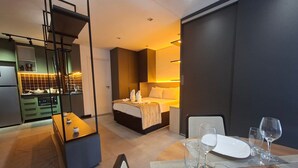 A modern hotel room with a kitchen area, a bed with a headboard, and a dining table set with glasses and plates. - Estúdios com piscina no lounge (São Paulo)