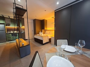 A modern hotel room with a kitchen area, a bed with a headboard, and a dining table set with glasses and plates.
