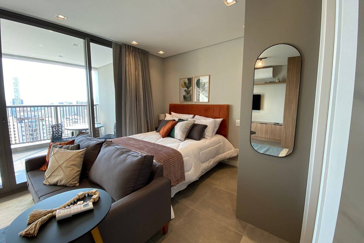 A modern hotel room with a large bed, a sofa, a small table, and a balcony with a view of the city.