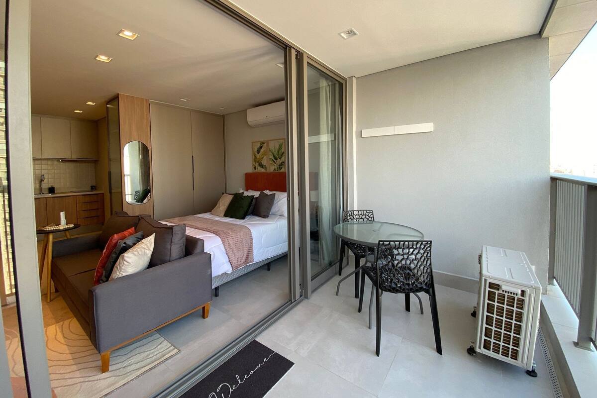 A modern hotel room with a bed, a sofa, a dining table, and a kitchen area visible through an open sliding door.