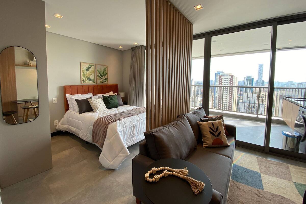 A modern hotel room with a bed, a sofa, a small table, a mirror, and a view of the city.