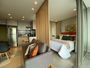 A modern hotel room with a kitchenette, a sofa with cushions, a bed with a headboard, and a sliding glass door.