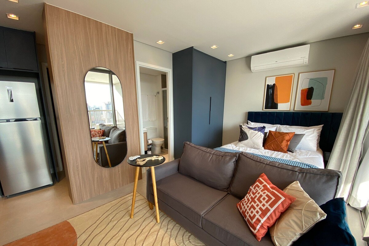 Una habitación de hotel moderna con sofá, una mesita, cama con almohadas, espejo y unidad de aire acondicionado.