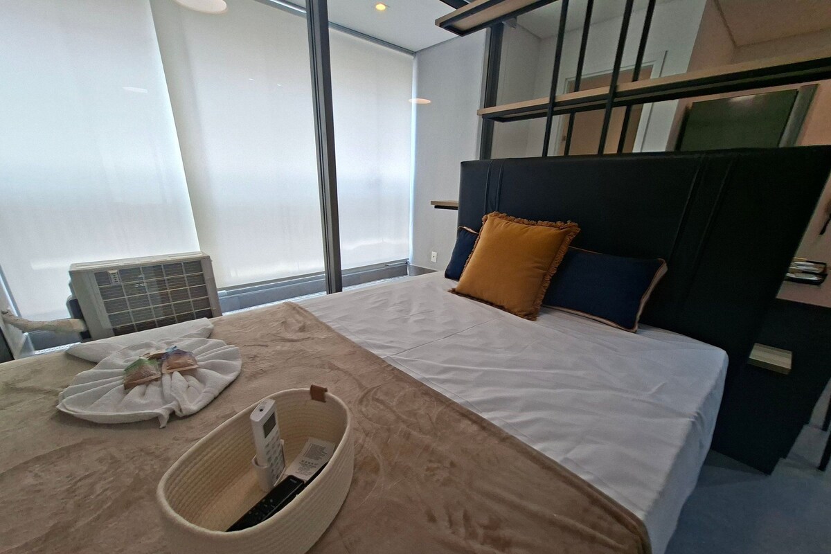 A hotel room with a bed, a headboard, a window, and a wall-mounted air conditioner.