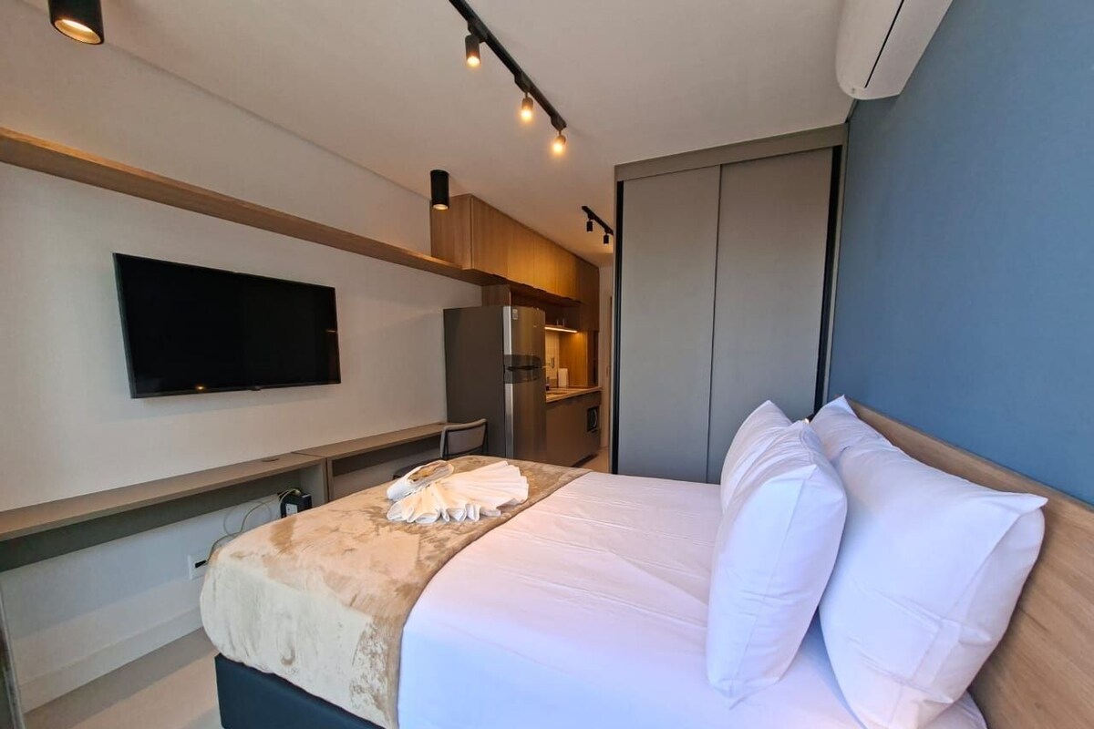 A hotel room with a bed, a television mounted on the wall, and a kitchenette area.