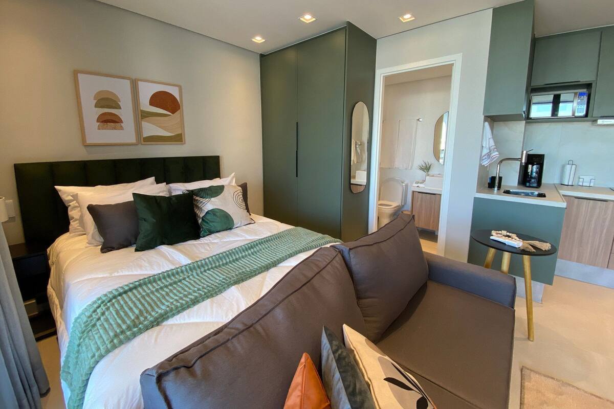 A modern hotel room with a bed, a sofa, a small table, a kitchenette, and a bathroom.