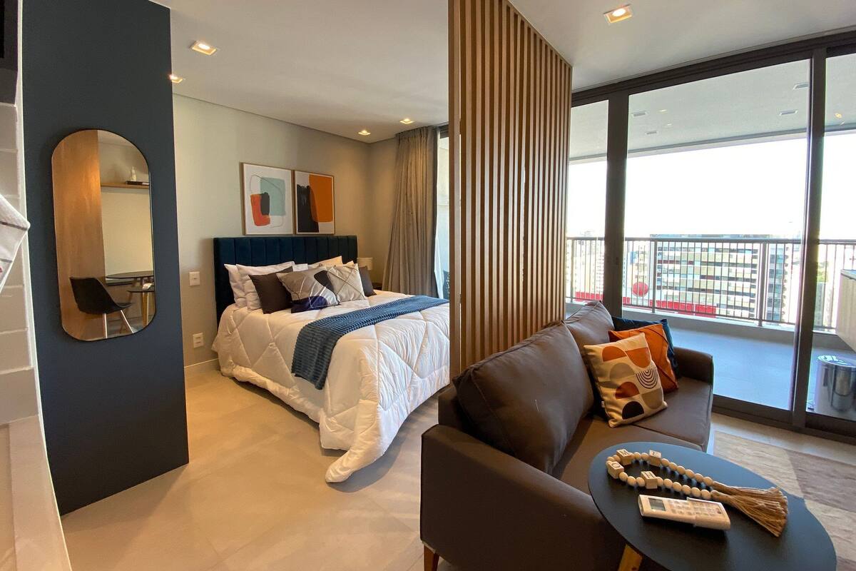 A modern hotel room with a bed, a sofa, a balcony with a view, and a large mirror.