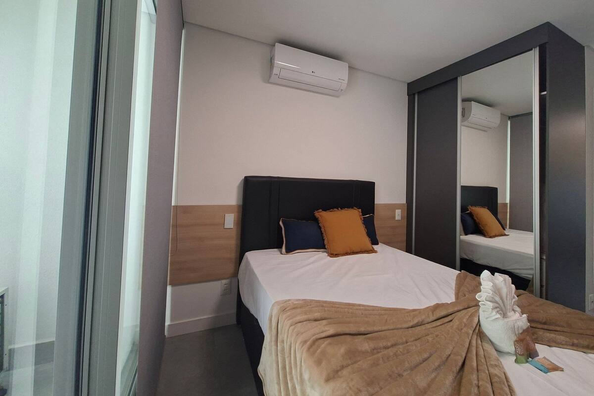 A hotel room with a bed, a large mirror, an air conditioning unit, and a window with curtains.