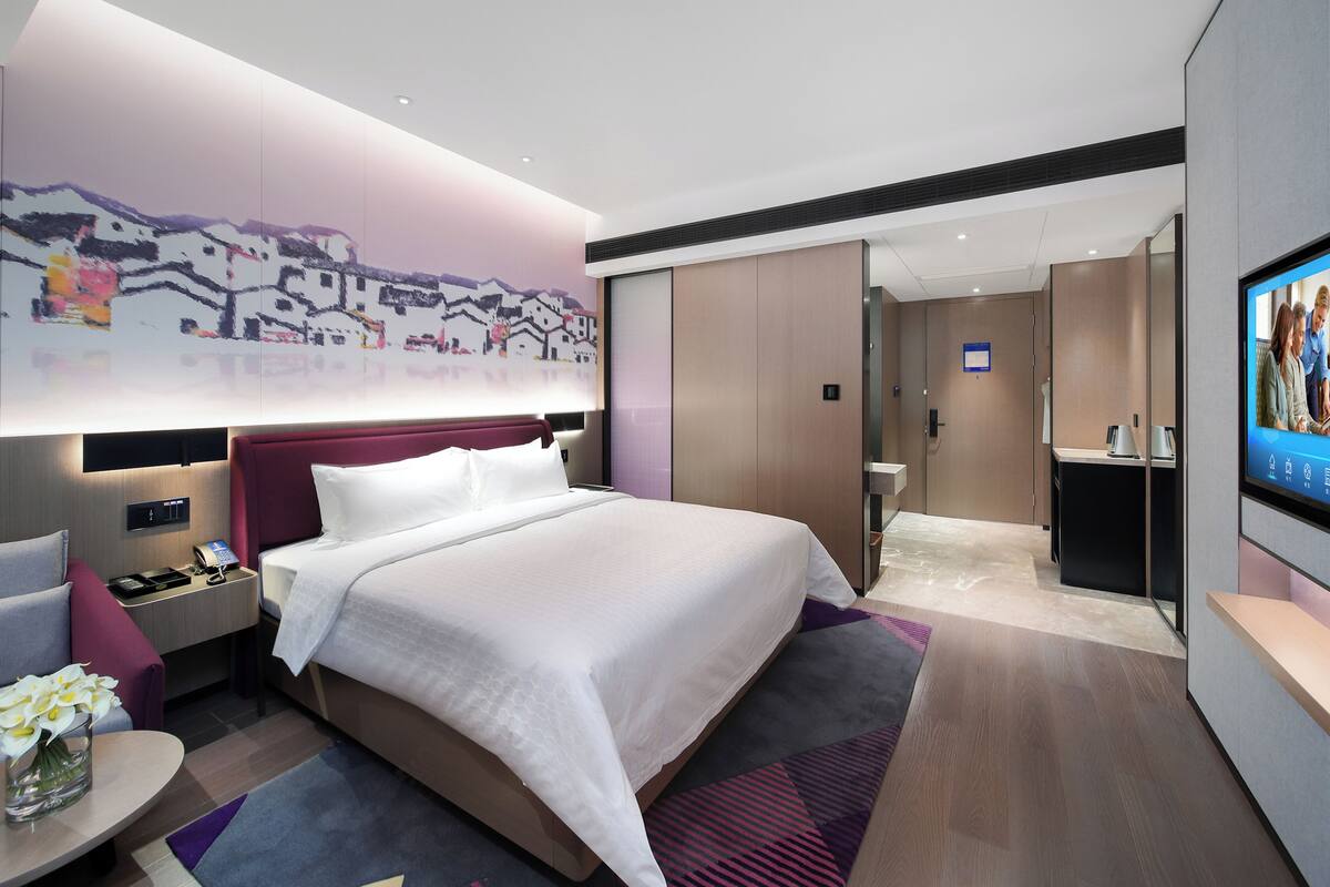 A modern hotel room with a large bed, a bedside table with a lamp, a TV, and a wall-mounted artwork.