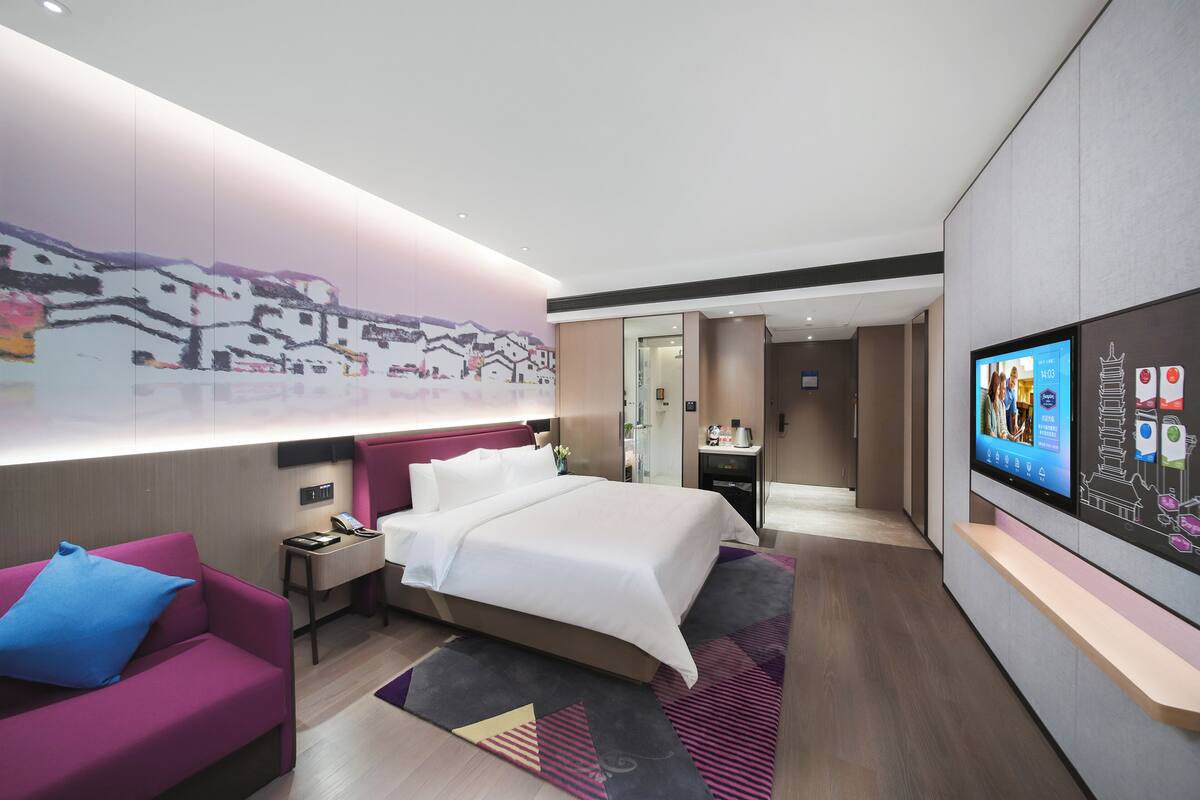 A modern hotel room with a large bed, a desk, a TV, and a wall-mounted artwork.