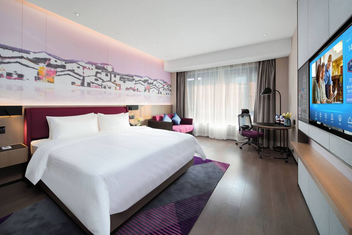 A modern hotel room with a large bed, a desk, a TV, and a cityscape mural on the wall.