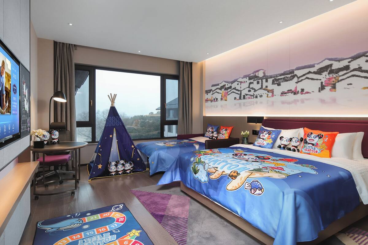 A hotel room with two beds, a desk, a chair, a TV, and a large window.