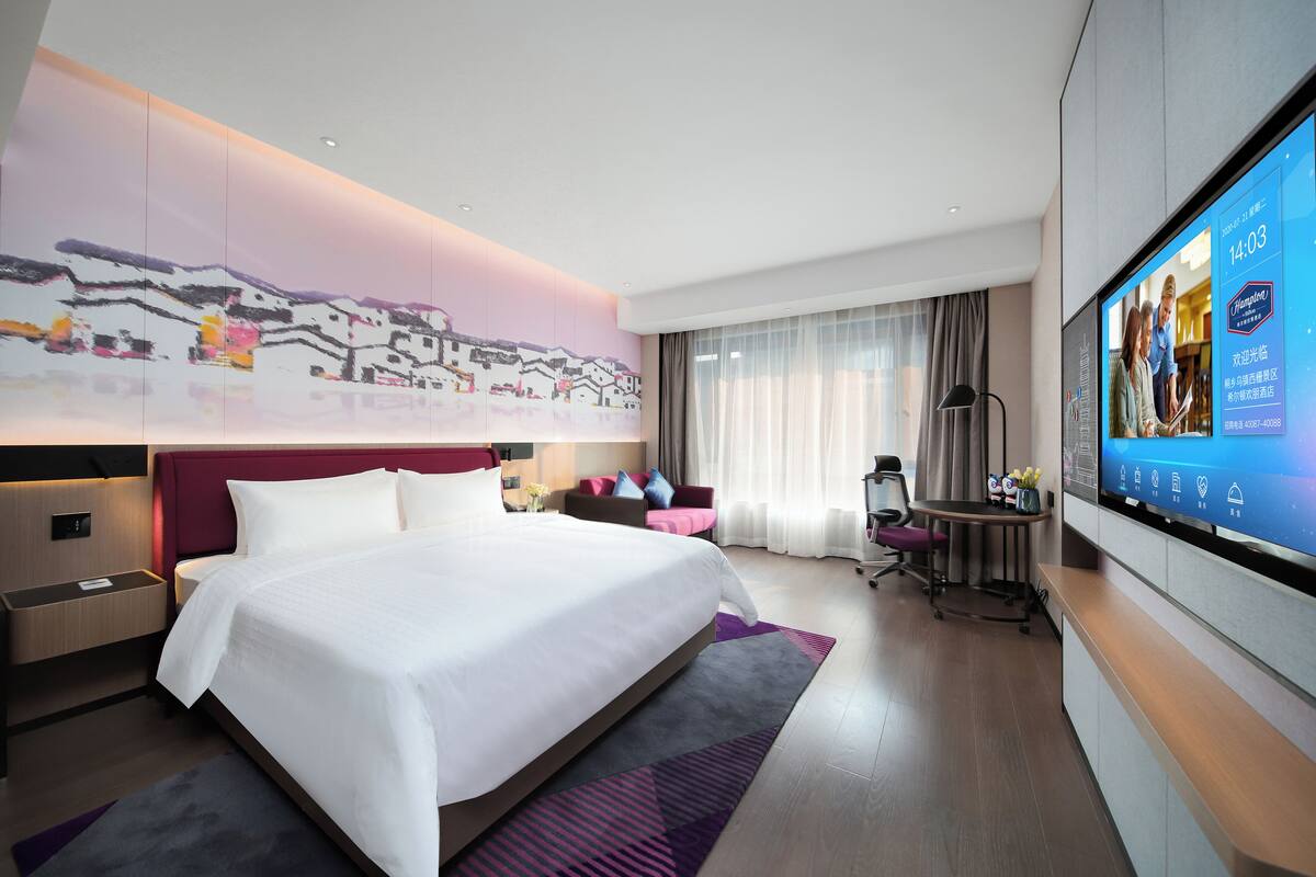 A modern hotel room with a large bed, a desk, a TV, and a view of the cityscape.