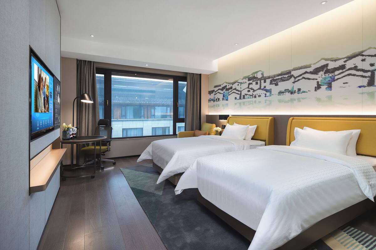 A hotel room with two beds, a desk with a computer, a TV, and a large window with a city view.