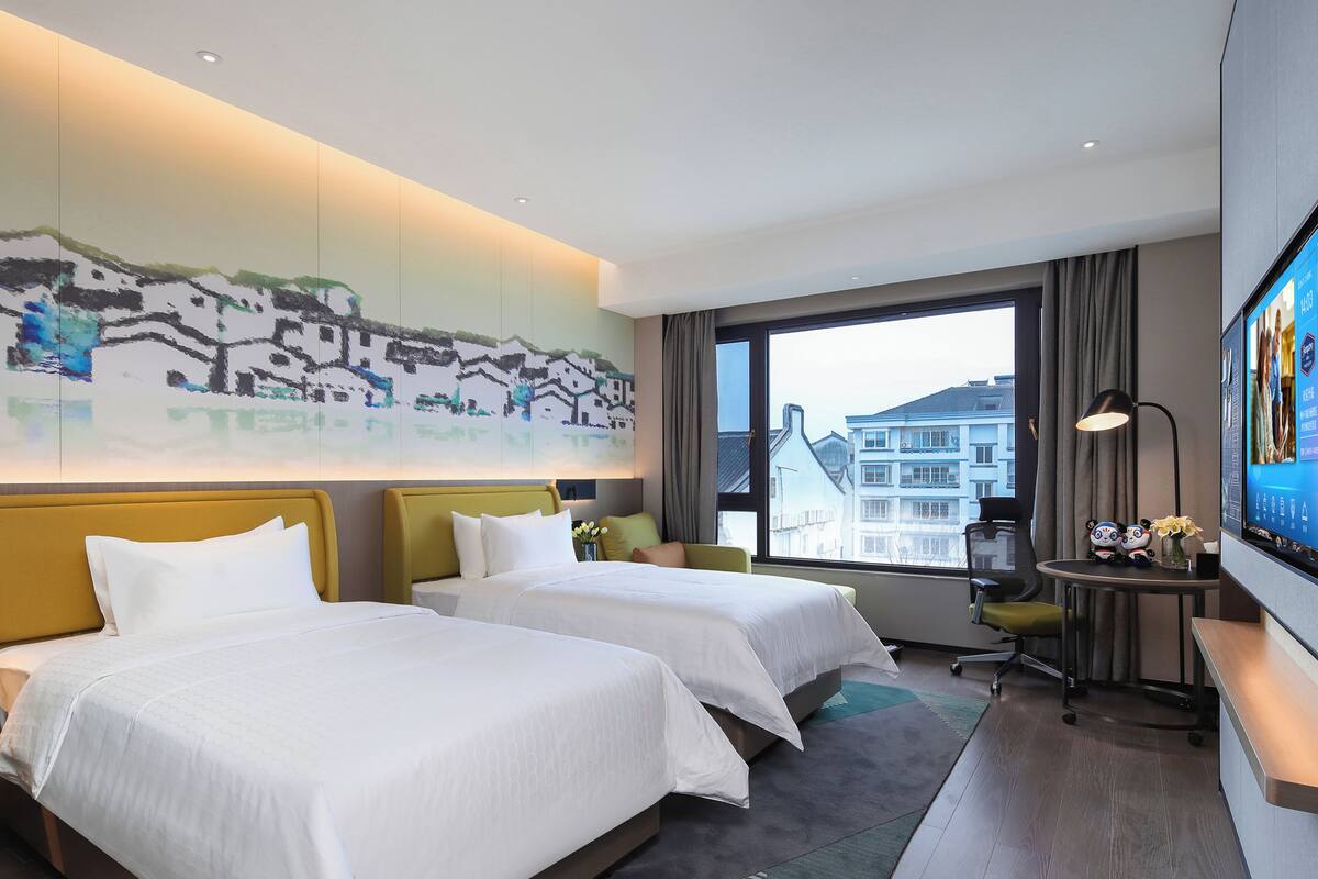 A hotel room with two beds, a desk with a computer, a TV, and a large window with a city view.