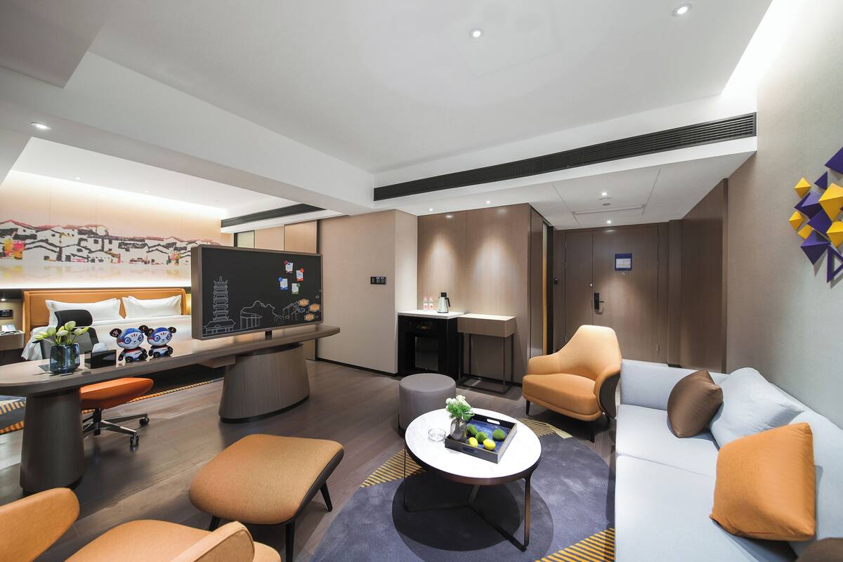A modern hotel room with a large bed, a desk with a computer, a sofa, and a coffee table.